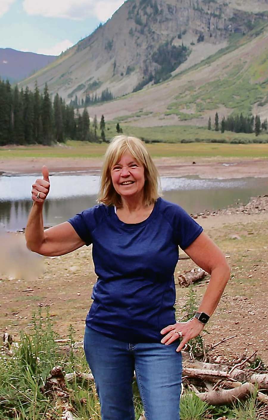Portrait photo of Suzanne Strauss showing a thumbs-up in front of a lake and mountain at a park