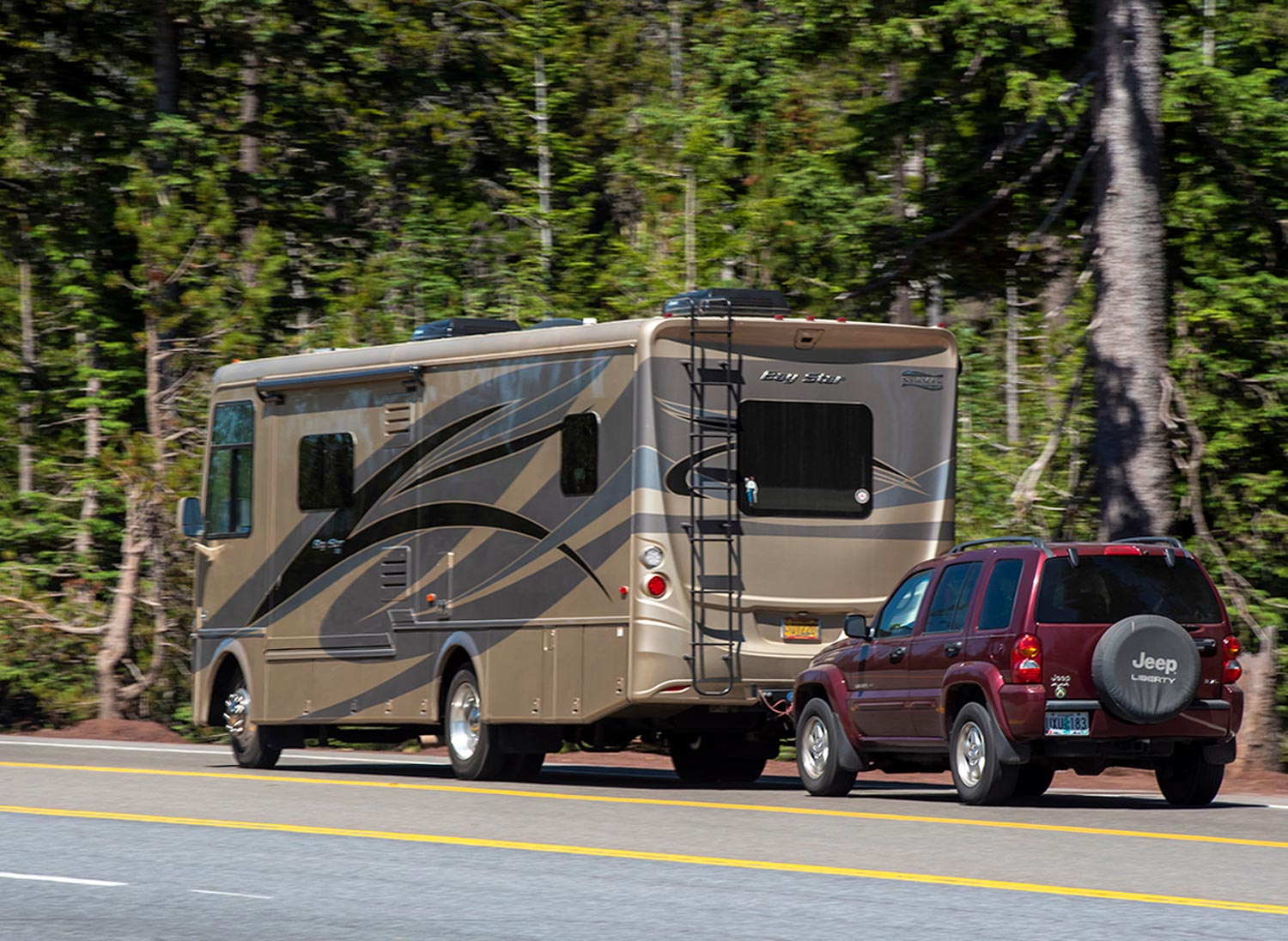 an RV towing an SUV down a road lined with trees