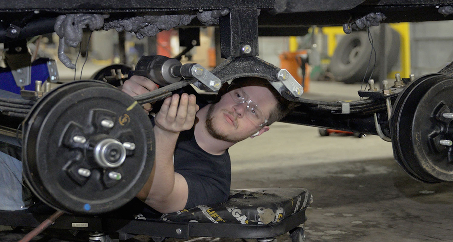 working under an RV, the mechanic uses a drill to unfasten all of the bolts from the stock equalizer and shackle brackets
