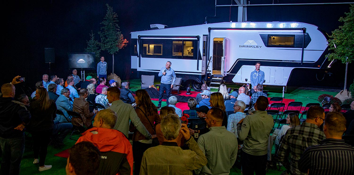 the Brinkley RV Model Z sits in full view as a large audience attending a promotional event listen to three spokesmen from Brinkley RV