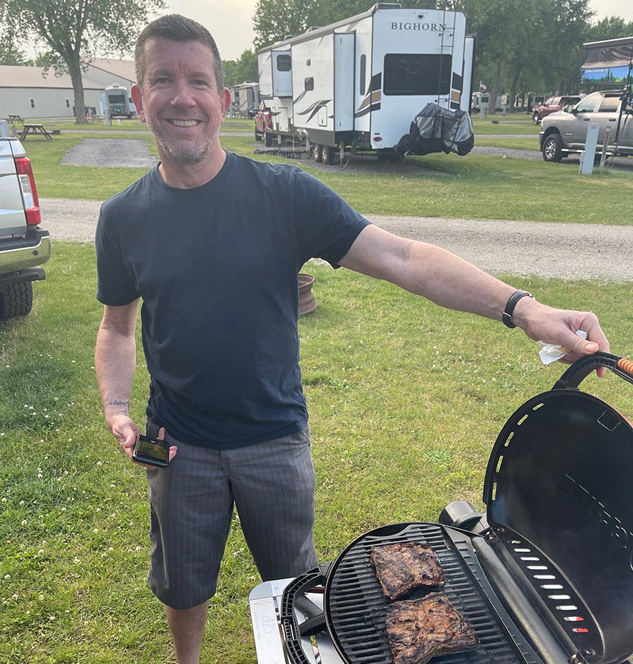 man smiling while grilling some steaks in front of an RV