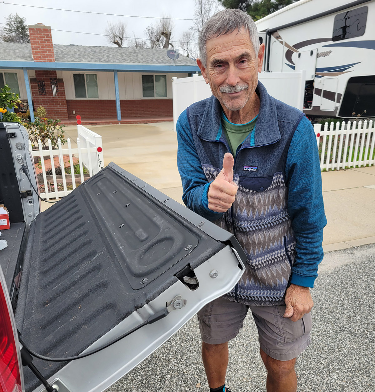 A portrait photograph of a man in a blue Patagonia jacket giving the thumbs up gesture grinning as the back bed/trunk door of the truck is open halfways on a gloomy cloudy day outside on a curb nearby a house and parked RV in the front yard