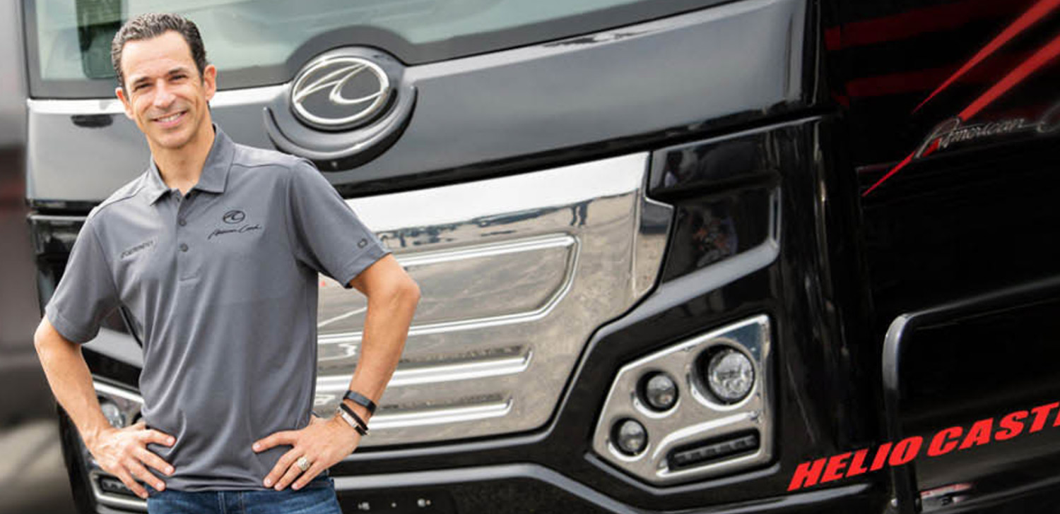 Hélio Castroneves smiles with his hands on his hips and wearing a polo shirt while standing in front of an American Coach RV