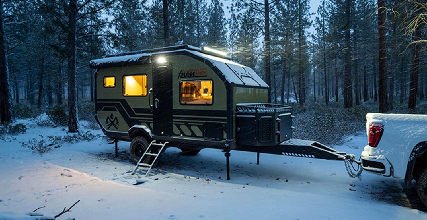 the 2023 Imperial Outdoors XploreRV X145 four-season overland trailer towed behind a parked white truck in a snowy forested area