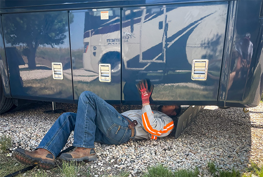 Exterior side photograph of a man quickly locating and repairing the source of the 2022 Newmar Ventana Class A's leaking air hose underneath the RV during the day