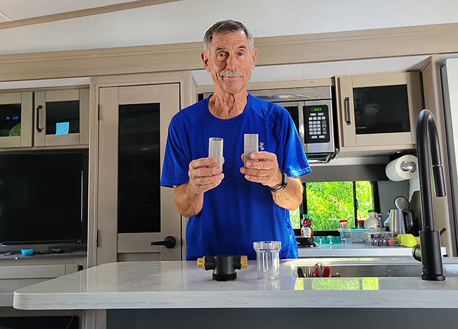Indoor RV close-up photograph perspective of Bill Gehr in a blue shirt holding small mesh screen filter tubes plus The Twinkle Star Garden Hose Filter Attachment product with the attachment's transparent jar tube are located on the marble countertop as he stands nearby the sink area