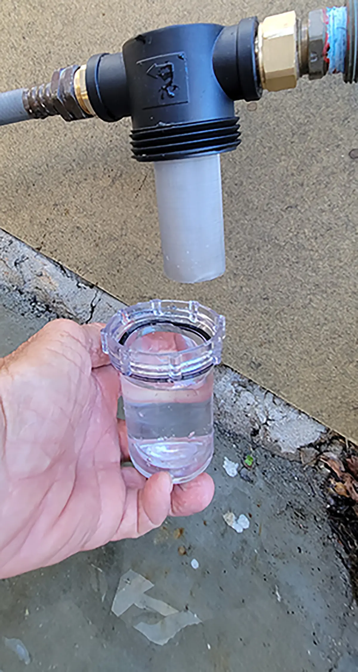 Close-up photograph perspective of Bill Gehr's hand holding a small transparent see through jar tube with water filled inside it and the mesh filter is attached to the head assembly located up above; 100 and 40 mesh filters are provided with the kit. The 100-mesh screen is equivalent to 149 microns. Cleaning the filter is simple; just remove the jar to access the mesh screen; it might require the use of a toothbrush or similar “tool” to removed stuck-on contaminants.