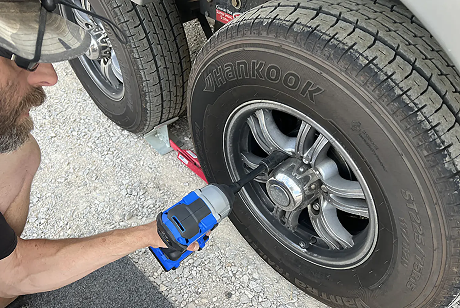Close-up photograph perspective of a man in sunglasses and a hat holding a big blue/grey power tool drill locking a solid lug-nut cap of/into a trailer wheel