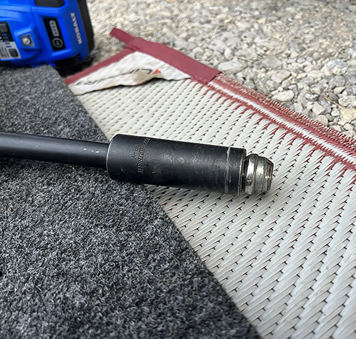 Close-up photograph perspective of a black metal original lug nut rod lied down on a grey mat next to a blue colored power drill tool
