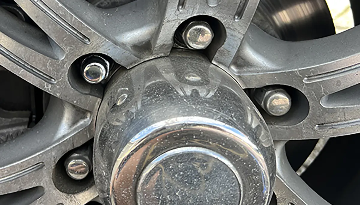 Close-up photograph perspective of the new lug nut (top left) is similar in style but a bit smaller than the original capped version (top right) of the trailer wheel; New lug nuts should be re-torqued after 50 miles, then again at 100 and 200 miles to assure proper seating.