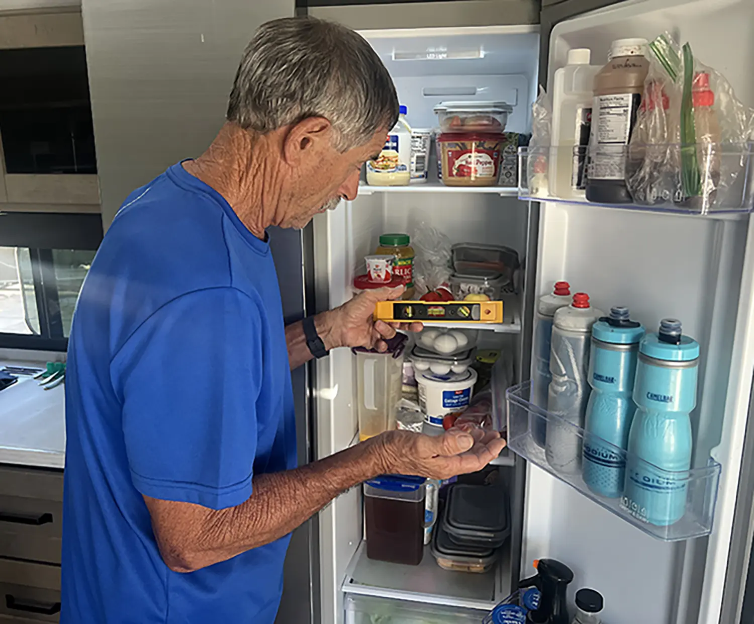 Indoor RV close-up photograph perspective of Bill Gehr in a blue shirt glancing downward at a yellow digital protractor device tool while holding the tool in his left hand as the refrigerator door is open