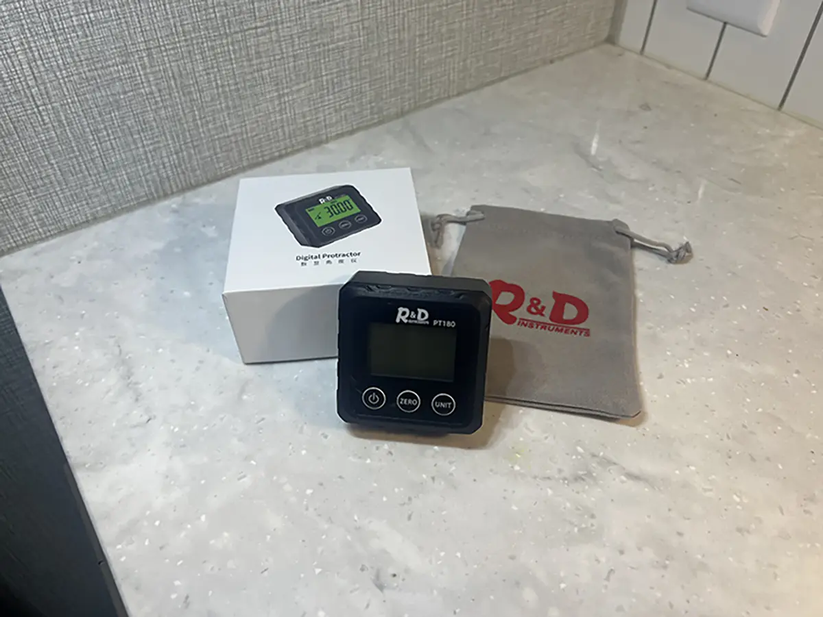Close-up photograph perspective of the Digital Angle Finder (Digital Protractor) from R&D Instruments on a white marble tile countertop; Also featured next to the device tool is a small dark grey storage bag with the R&D Instruments printed logo on the storage bag and the small white product box of the device tool situated across from the storage bag