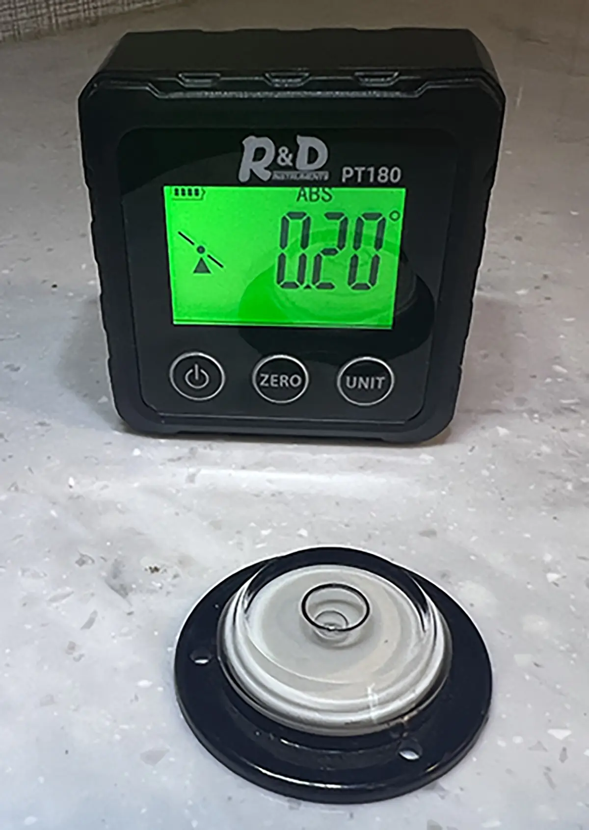 Close-up photograph perspective of the Digital Angle Finder (Digital Protractor) screen reading (in green) from R&D Instruments on a white marble tile countertop; The reading shows ABS 0.20 degrees, a fully charged battery icon, and a slant tilted icon symbol while in front of the digital protractor device device tool is a round circular bubble object with a round covering lid