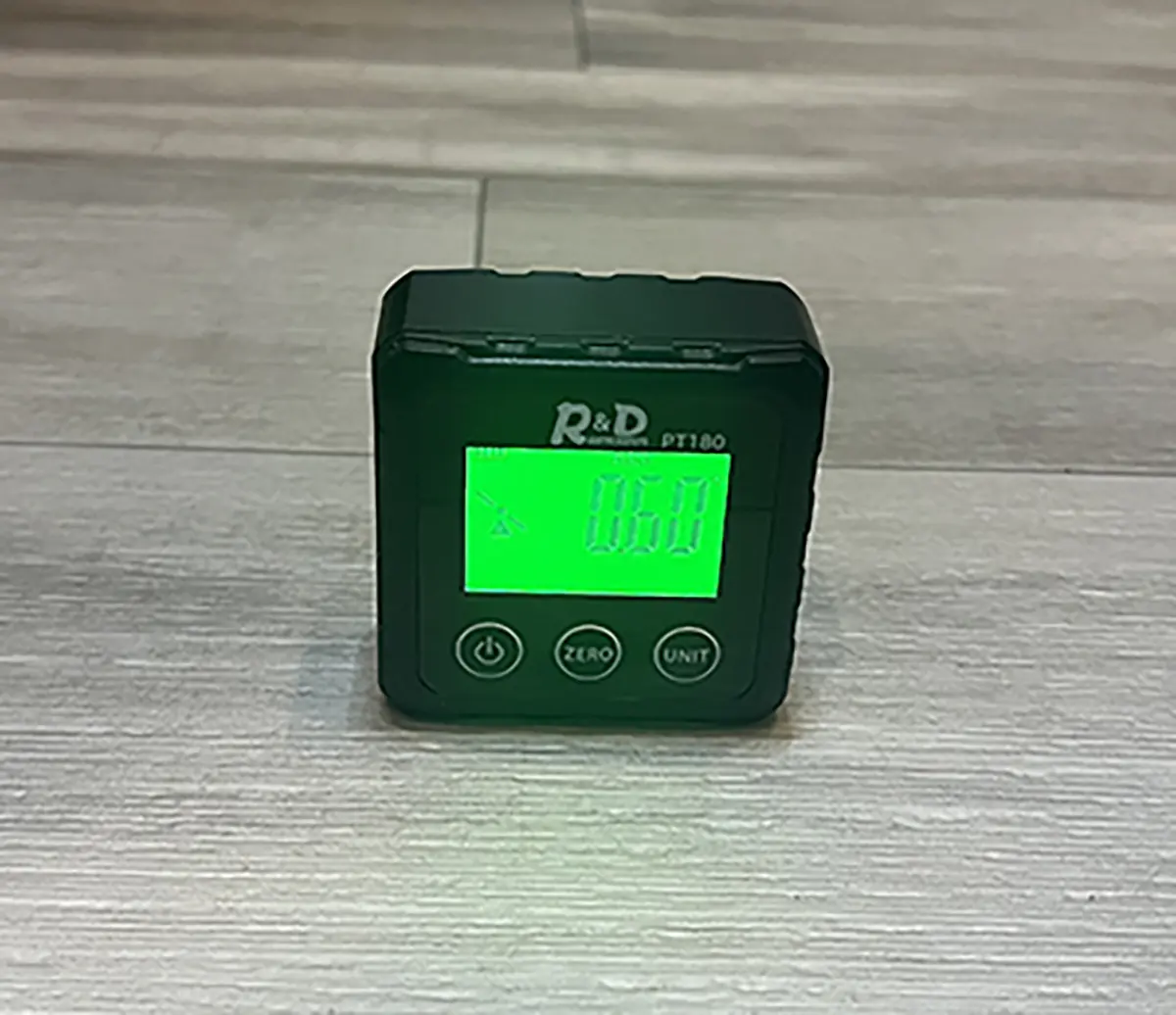 Close-up photograph perspective of the Digital Angle Finder (Digital Protractor) screen reading (in green) from R&D Instruments on a dark grey tile countertop; The reading shows 0.60 degrees and a slant tilted icon symbol