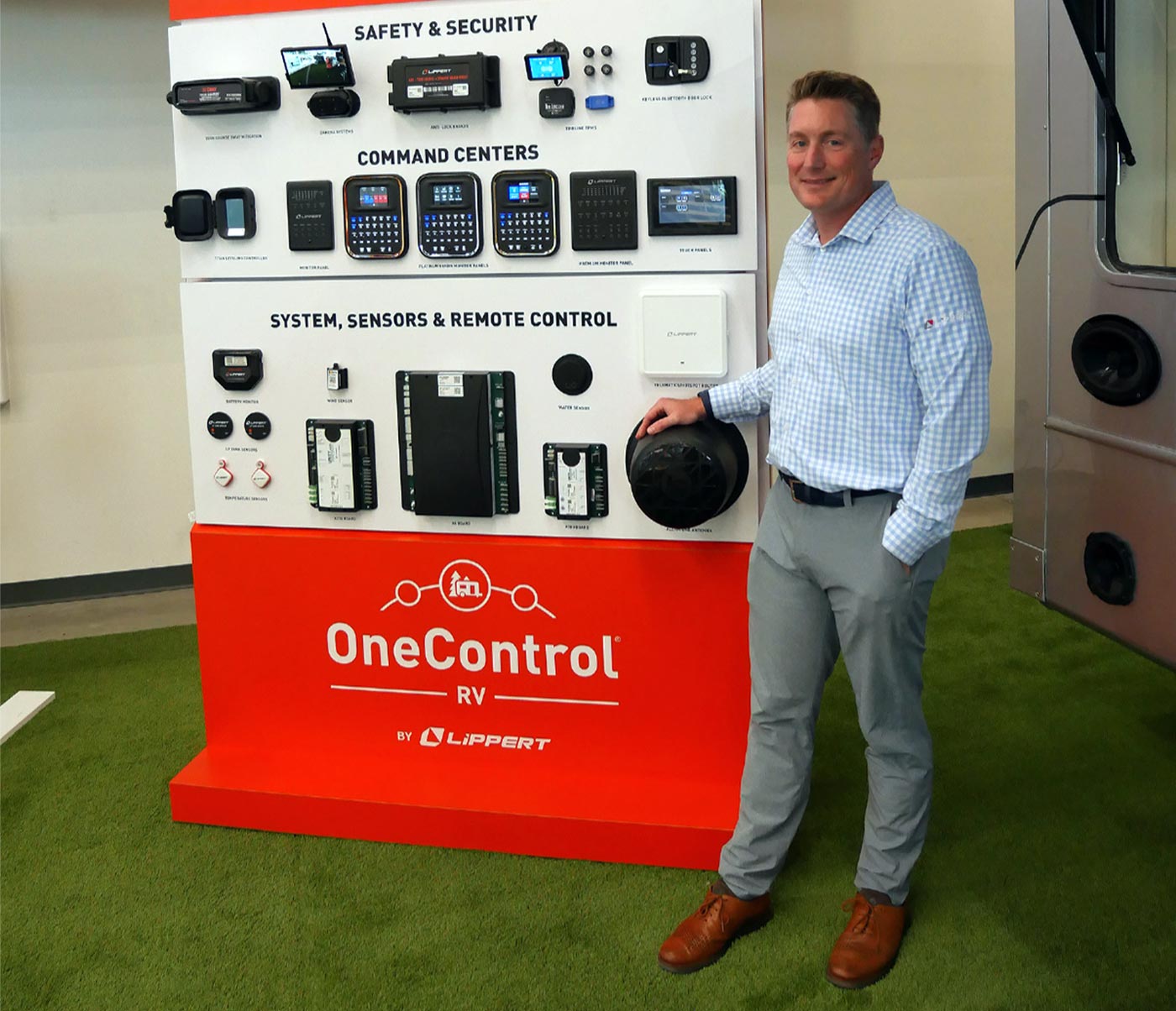 Jarod Lippert wearing a light blue plaid button up shirt and slacks smiles while standing alongside a OneControl display showing the wealth of functions the device now controls