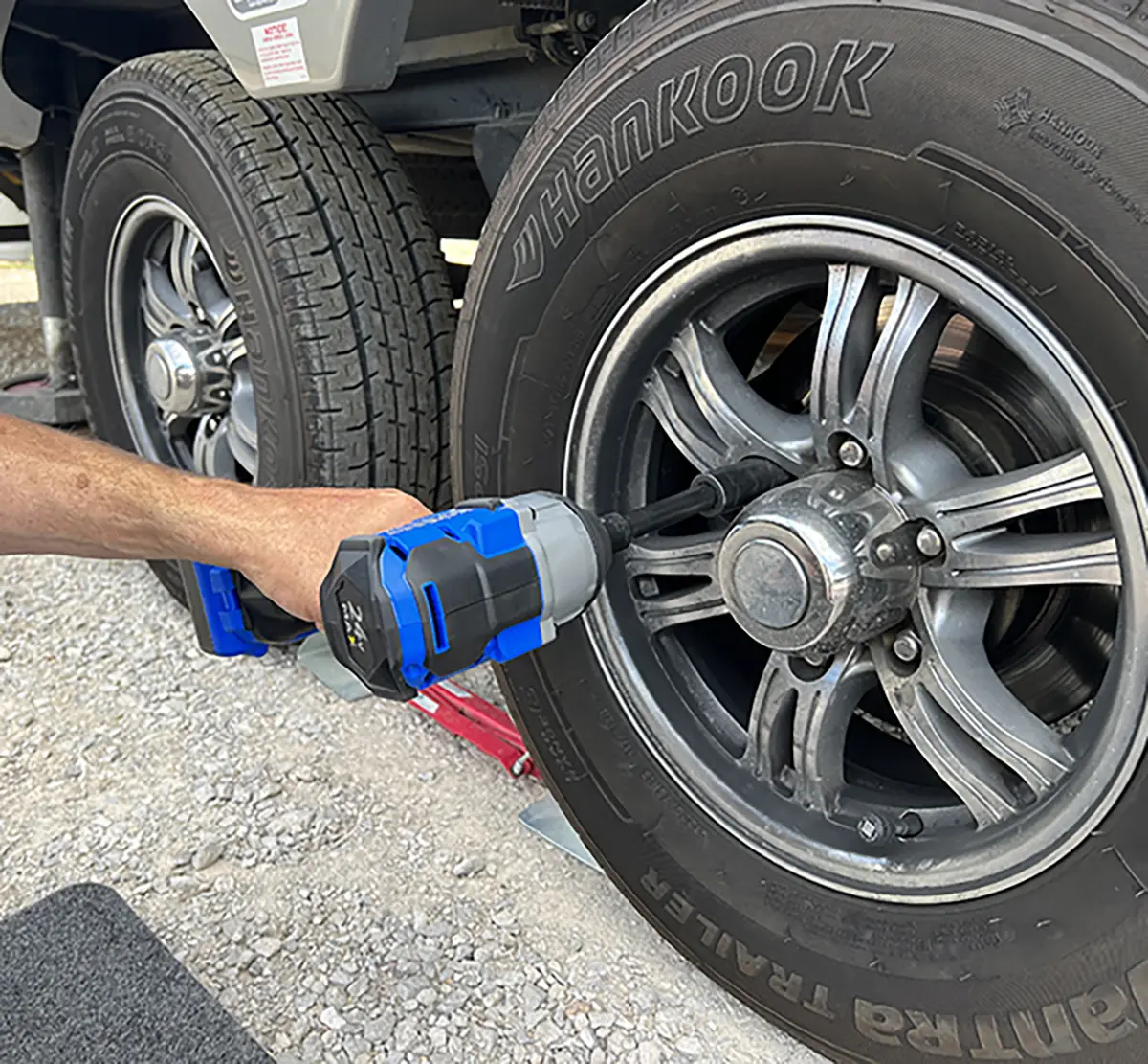 Close-up photograph of a man's hand using the blue/grey power drill too on the trailer wheel after threading a new lug nut on to the stud, using a socket and extension, to prevent cross threading