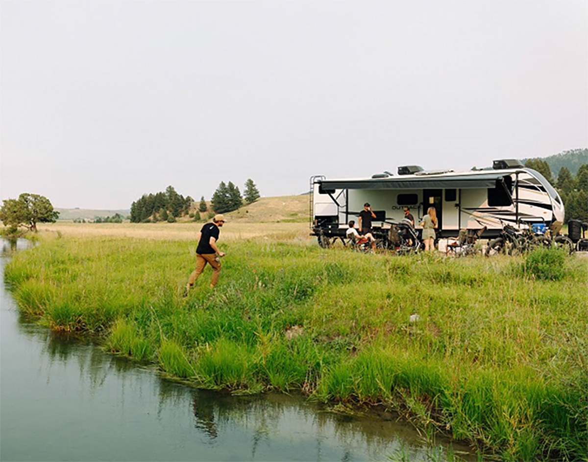 Man walking away from a pond and toward an RV that is parked in knee high grass, a few trees in the distance