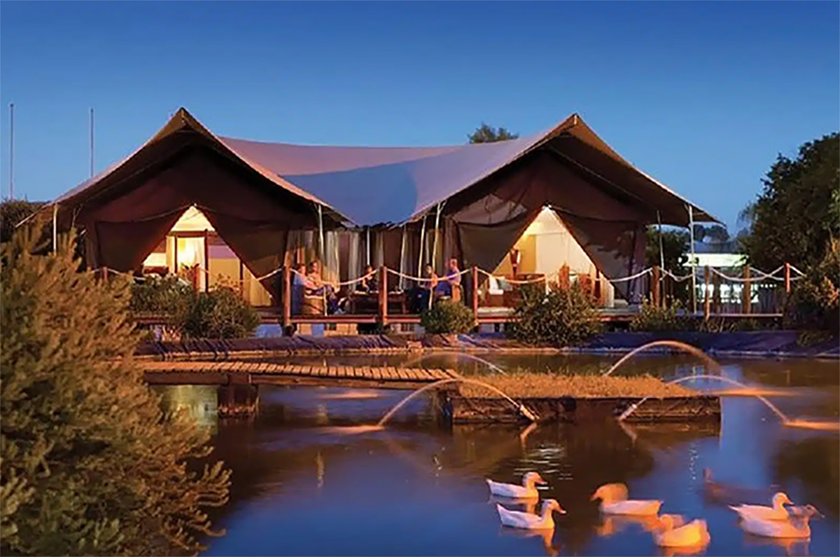 Large glamping tent in the evening, with lights on inside, situated behind a pond with ducks in it