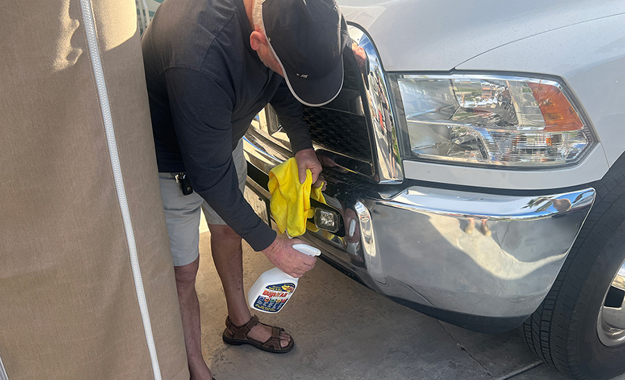 Landscape close-up photo view of a man in a black hat, black long sleeve t-shirt, light grey shorts, and strapped cameltoe sandals holding a ProSol Bugs N All Multi-Use Vehicle Cleaner spray bottle product model in his hands as he sprays the liquid into the front bumper area of his white truck while holding a yellow rag towel in his other hand