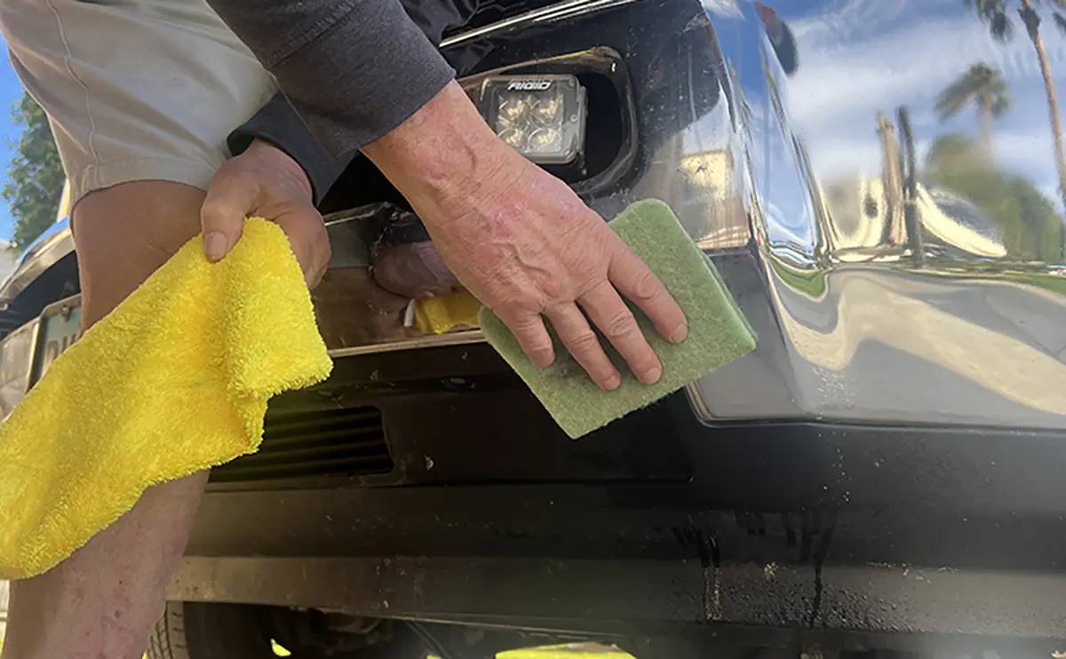 Landscape close-up photo view of a man's hand holding a yellow rag towel in one of his hands and using the light forest green Bugs Off Pad product with his other hand on his truck's bumper