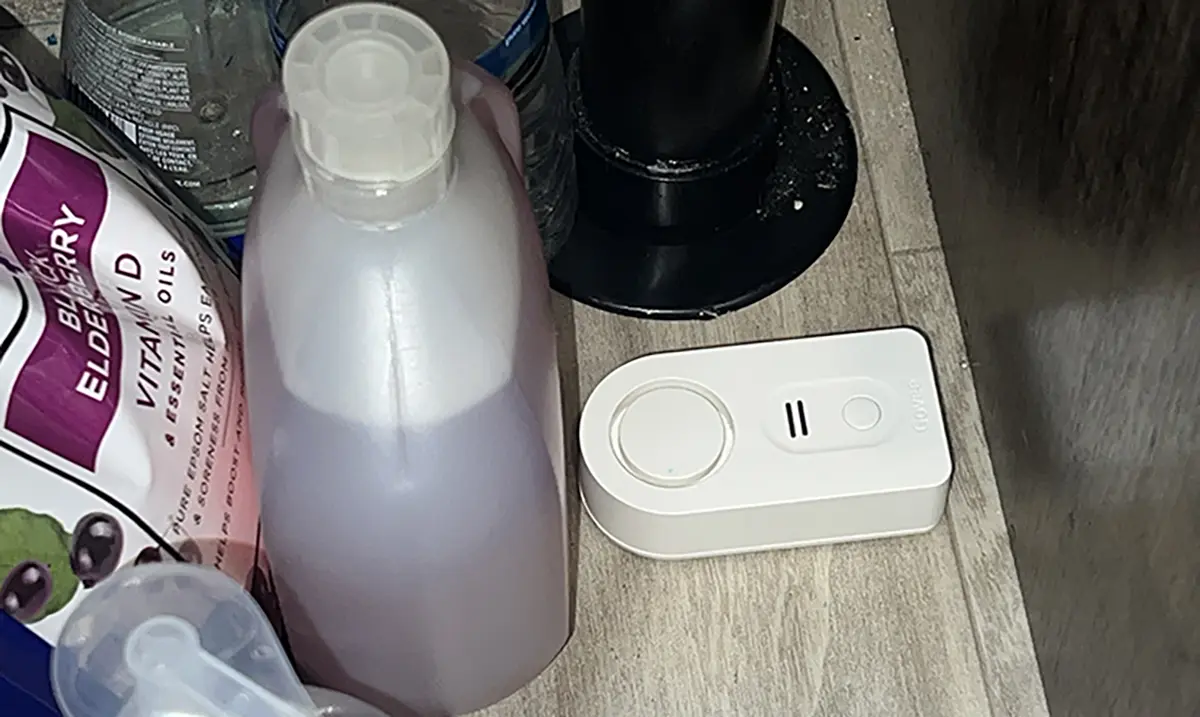 Landscape close-up photo view of the Govee H5054 water alarm sensor product model placed on the floor situated nearby some other random assorted RV cleaning products inside a RV kitchen setting area