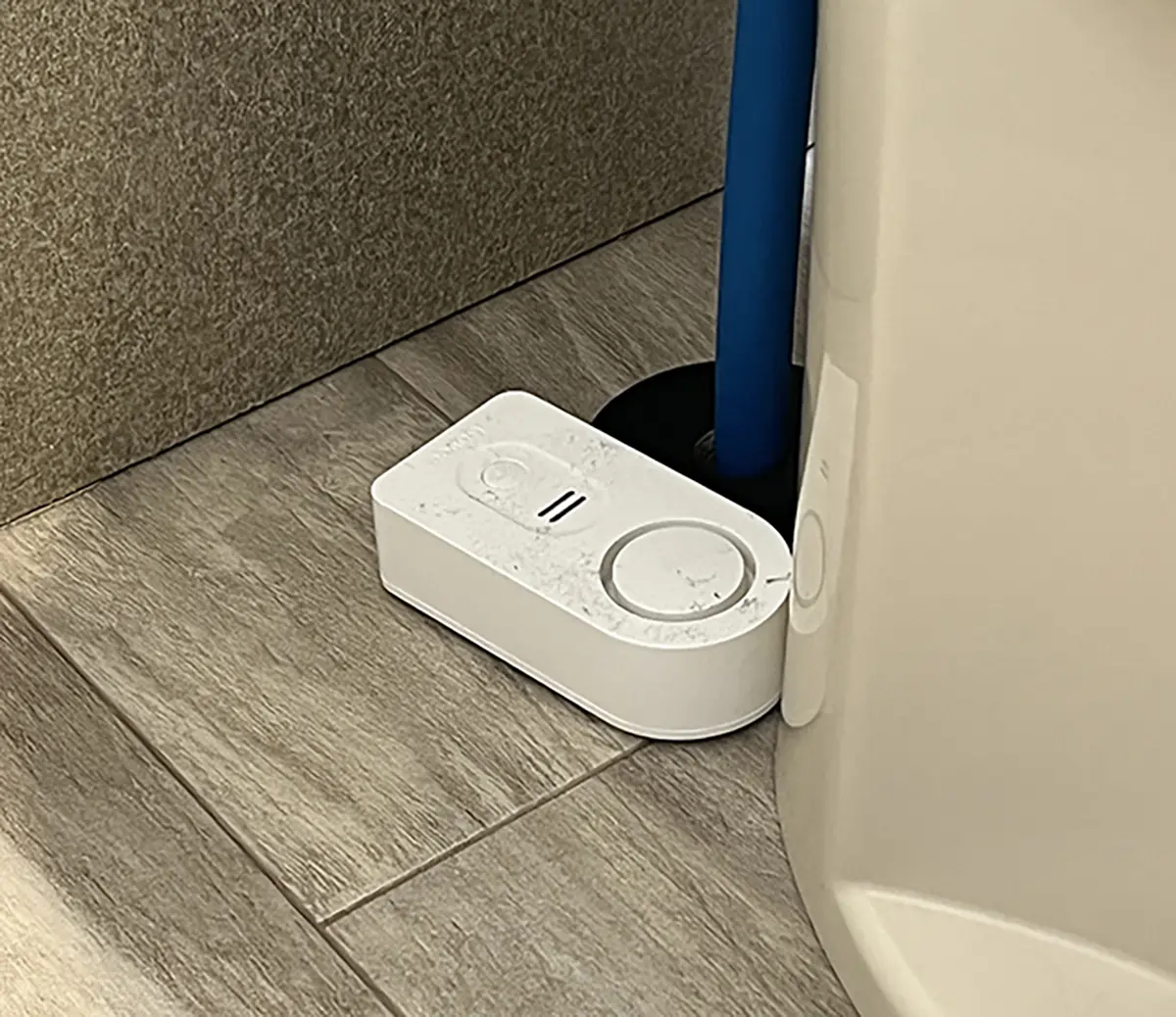 Portrait close-up photo view of the Govee H5054 water alarm sensor product model placed on the floor situated nearby a toilet inside a RV bathroom setting area