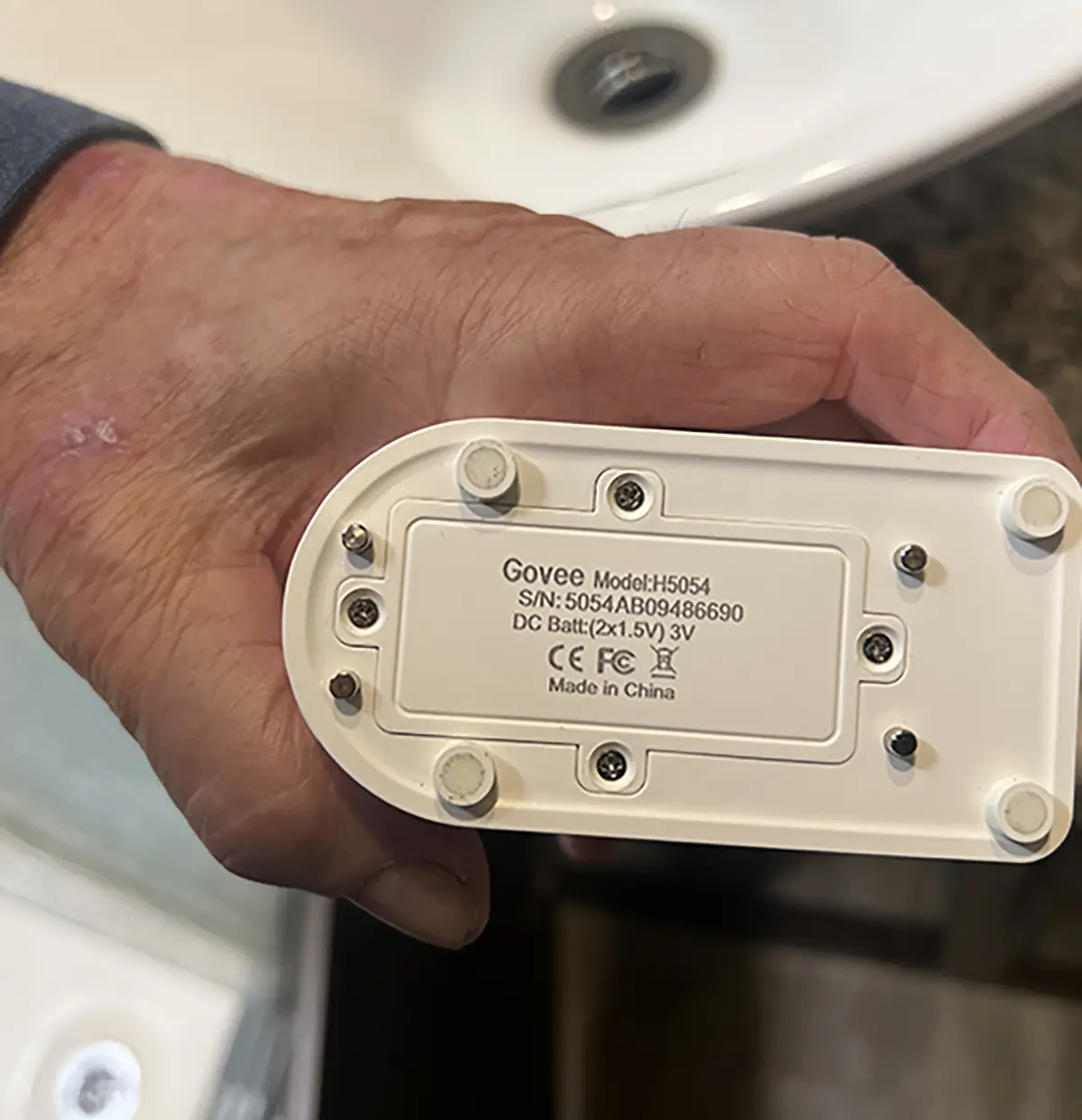 Portrait close-up photo rear back view of a man's fingers holding a Govee H5054 water alarm sensor product model in his hand inside a RV setting area as the label description details are shown such as the name of the model, S/N number and/or letter combination, DC Battery type, etc.