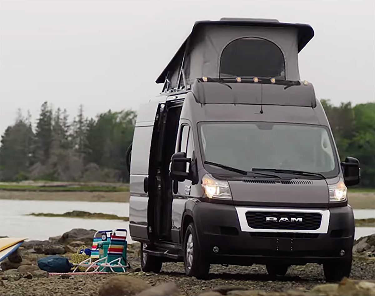 Outdoor portrait close-up photo view of a black Dodge Ram Promaster Class B RV van vehicle parked on some sand soil ground with its back rear right side passenger sliding door open and an equipped shaded canopy cover accessory attached on top of the vehicle as there are beach lawn chairs, lush green trees, and a body of water nearby in the background on a gloomy overcast day