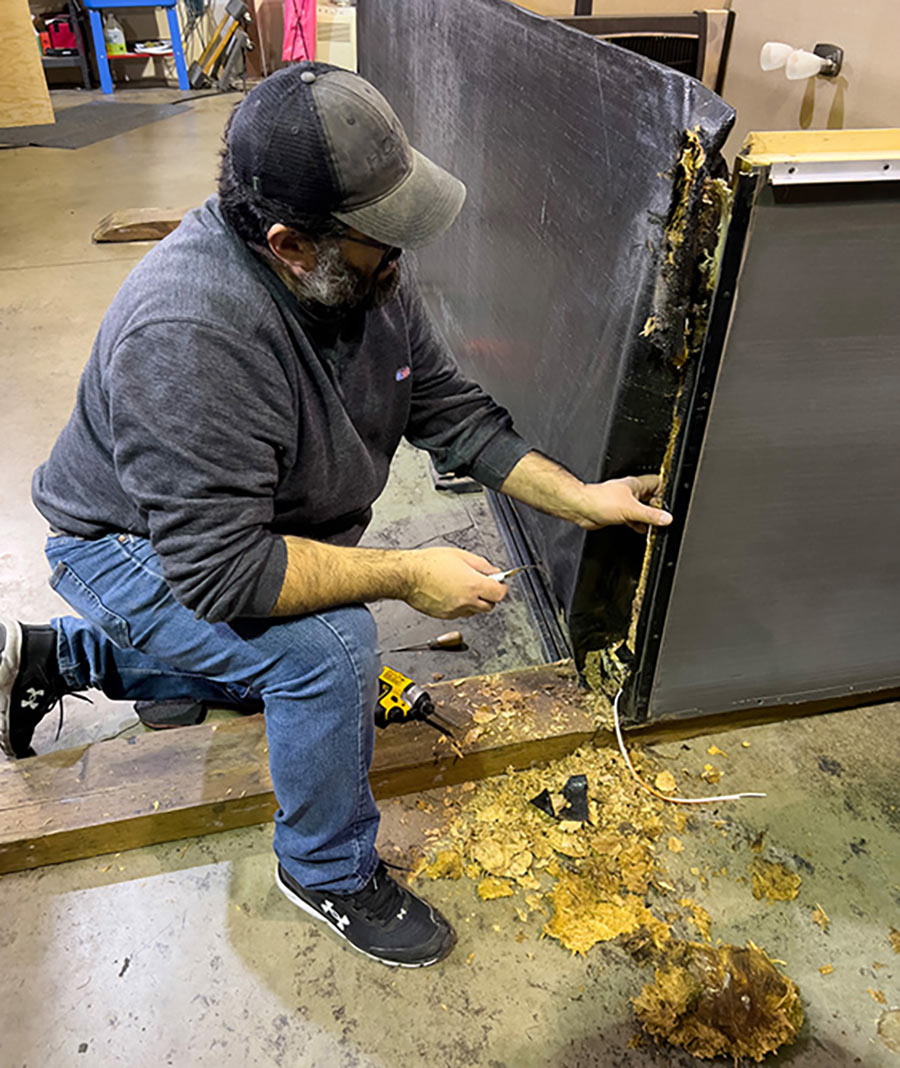 Portrait close-up indoor photograph of a man in a faded dark black/grey hat, dark faded navy blue long sleeve t-shirt, light blue denim jeans, and black Under Armour fitness shoes kneeling down on the ground glancing at wood in a workshop environment area as the OSB flooring used in most of the slides in today’s RVs is very susceptible to rot when it comes into prolonged contact with water