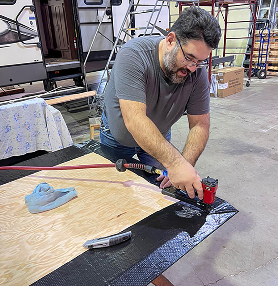 Portrait close-up photo view of a man in a dark grey t-shirt and light blue denim jeans using a tool to glue and staple all the edges and folds around the top of the new wooden RV slide board floor as it doesn’t require full vapor barrier coverage because it is inside and will be covered by carpet or other flooring material; However, it still needs to be very secure to keep the vapor wrap tight.