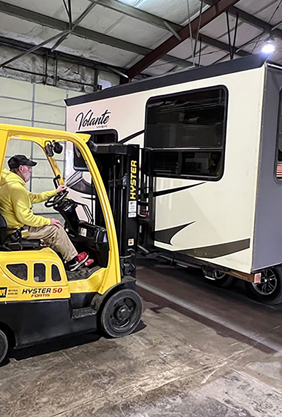 Portrait close-up photo view of a man in a yellow sweater, black hat, light beige pants, and red/black Nike fitness shoes driving a forklift as it latches onto the RV trailer; Having a forklift makes the job of removing and re-installing a slide less complicated. Take reinstallation very slow so the seals aren’t damaged.
