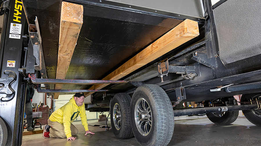 Landscape close-up photo view of a man in a yellow sweater, black hat, light beige khaki pants, and red/black Nike fitness shoes glancing across a particular RV sub-floor replacement, which was installed with 10-foot-long 4x4s under the slide so it can be lifted by a forklift; Once the weight of the slide is on the forks, it’s unbolted from the two rams and carefully pulled out of the trailer.