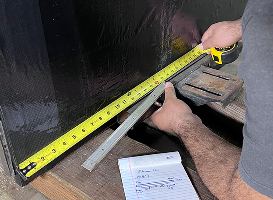 Landscape close-up photo view of a man's hands/arms measuring a piece of wood with a measuring tape; Before anything is removed from the slide, one thing of note is the exact measurements of the slide ram mounting bracket that runs the width of the room so it can be bolted back in the same location as it came from the factory. This eases repositioning of the slideout so it fits properly in the cutout.