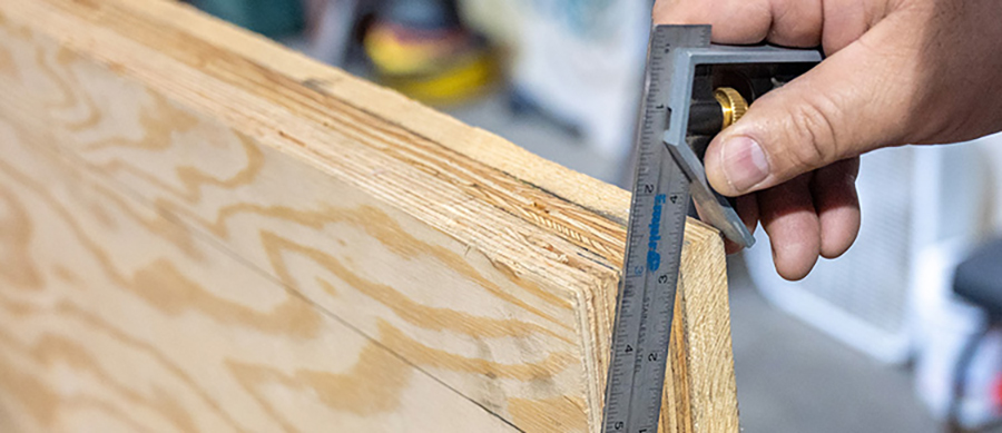 Landscape indoor close-up photo view of a man's fingers holding a one-inch-thick marine-grade plywood near a layer of wood, which plywood is ideal for use as a replacement sub-floor in RV slides; This type of plywood is highly resistant to water intrusion, and provides a stronger, more rigid floor. Transfer the taper measurements to the replacement floor. The extra ½-inch wood pieces shown here serve as a guide for the router that will be used to cut the angle on this 14-foot-long sheet of 1-inch marine-grade plywood.