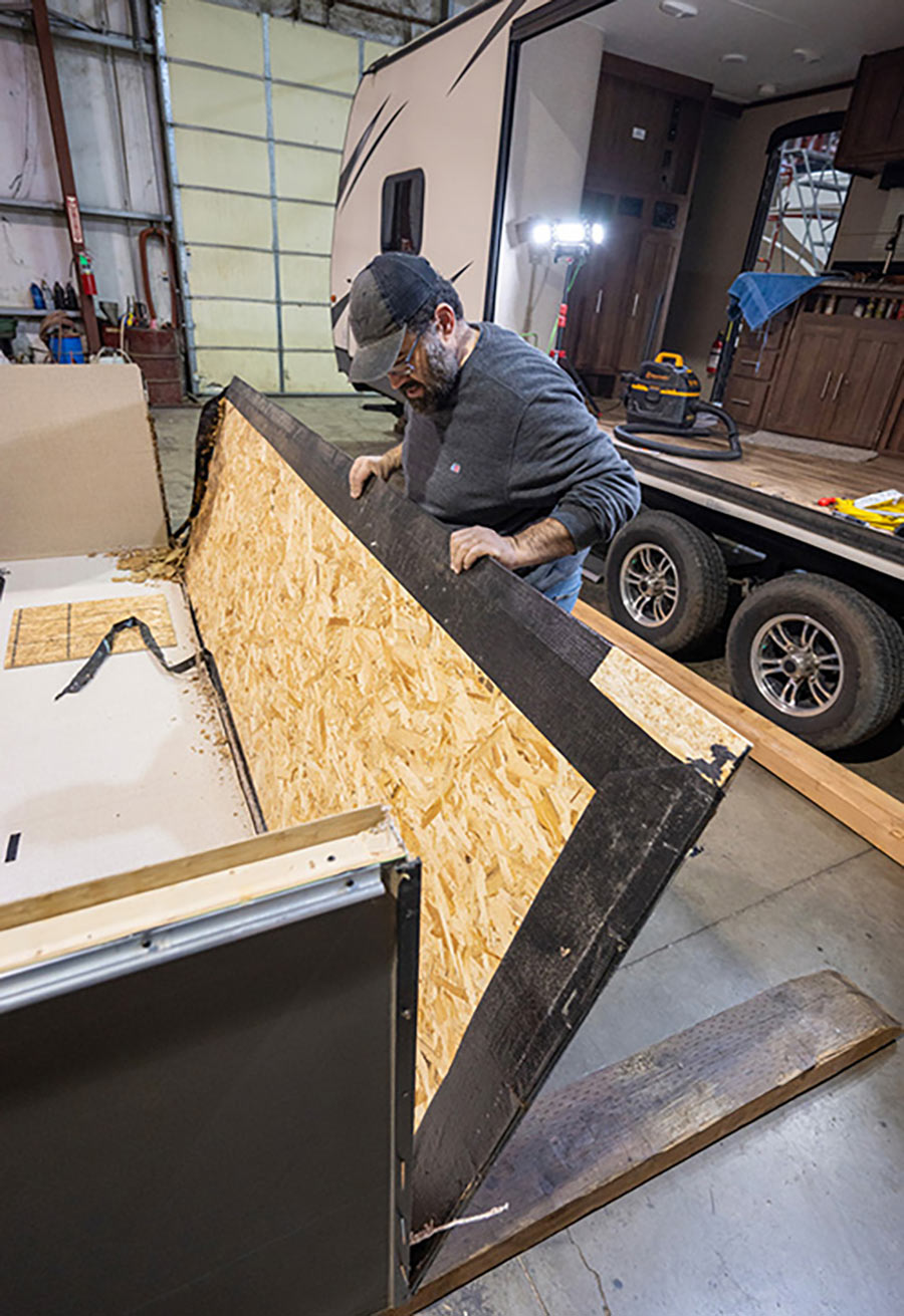 Landscape close-up photo view of a man in a dark black/grey hat and faded dark navy blue long-sleeve t-shirt glancing down at a partial rotted wooden floor board as it is lifted upwards halfways in the air