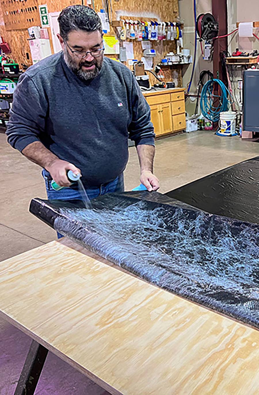 Close-up indoor portrait photo view of a man in a dark faded navy blue long-sleeve t-shirt and light blue denim jeans using a 3M 90 Hi-Strength Spray Adhesive onto a sheet of custom-made inch-thick marine-grade plywood acting as a quality vapor barrier material being laid out onto the new wooden RV slide floor board; This job required two cans of the adhesive.