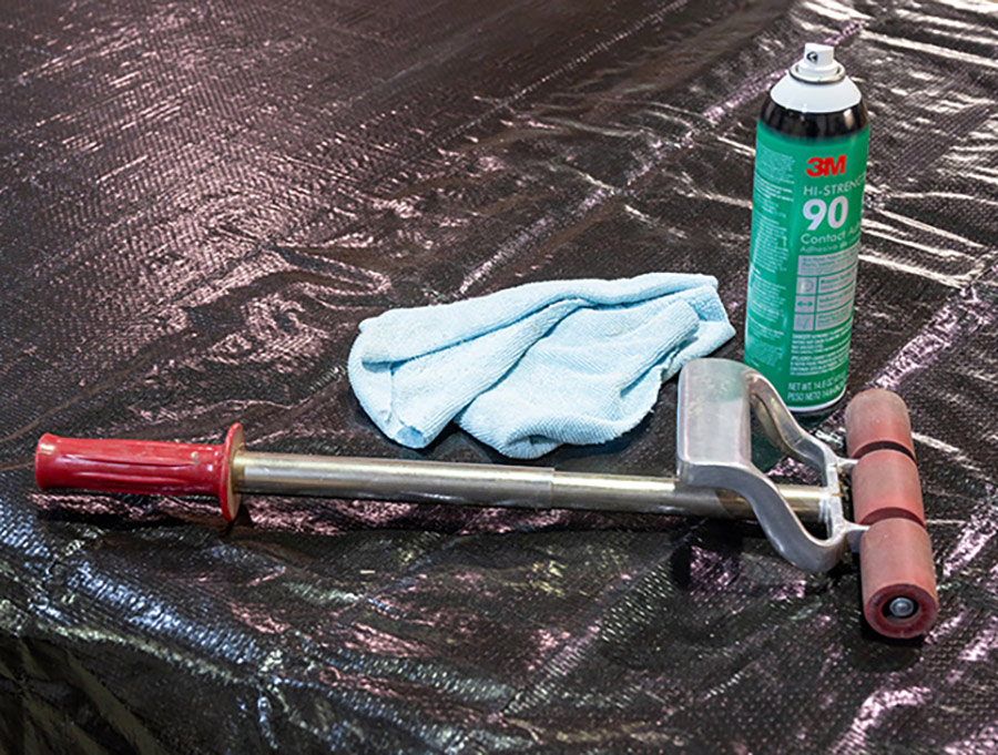 Close-up landscape photo view of a 3M 90 Hi-Strength Spray Adhesive canister, a light sky blue rag towel, and a heavy duty roller object displayed on top of a black cover material