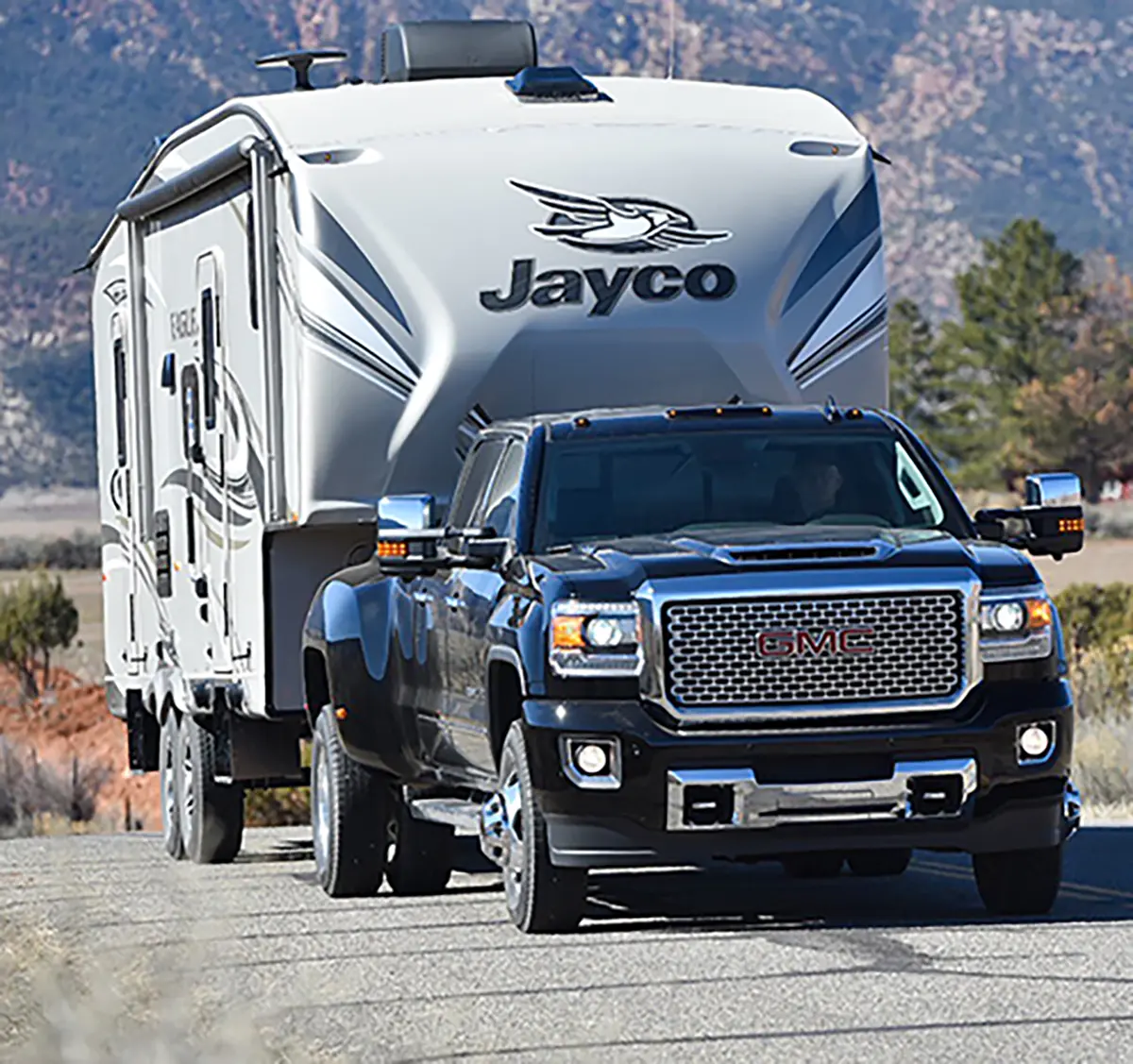 Close-up portrait outdoor photograph view of a man seated inside a GMC truck model driving down the road hauling a Jayco RV trailer behind on the GMC truck's hitch during the sunny bright day