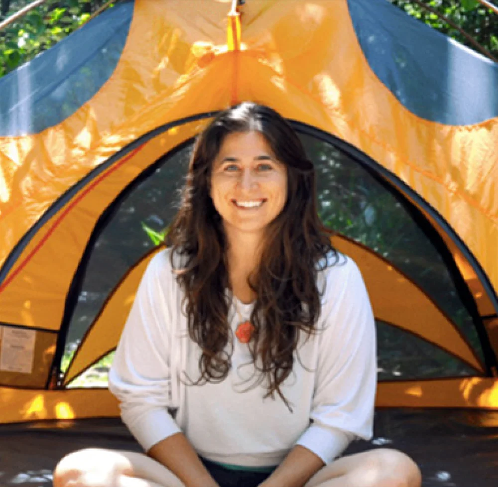 Alyssa Ravasio, a smiling woman with long brown hair, sits cross legged in front of an orange camping tent