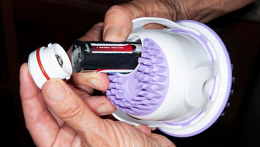 Close-up portrait photograph view of hands feeling/holding the Orb Spa Vibra sonic massaging option with the battery popped out, in which the motor that creates the vibrations operates on three AAA batteries, which are placed in a waterproof compartment