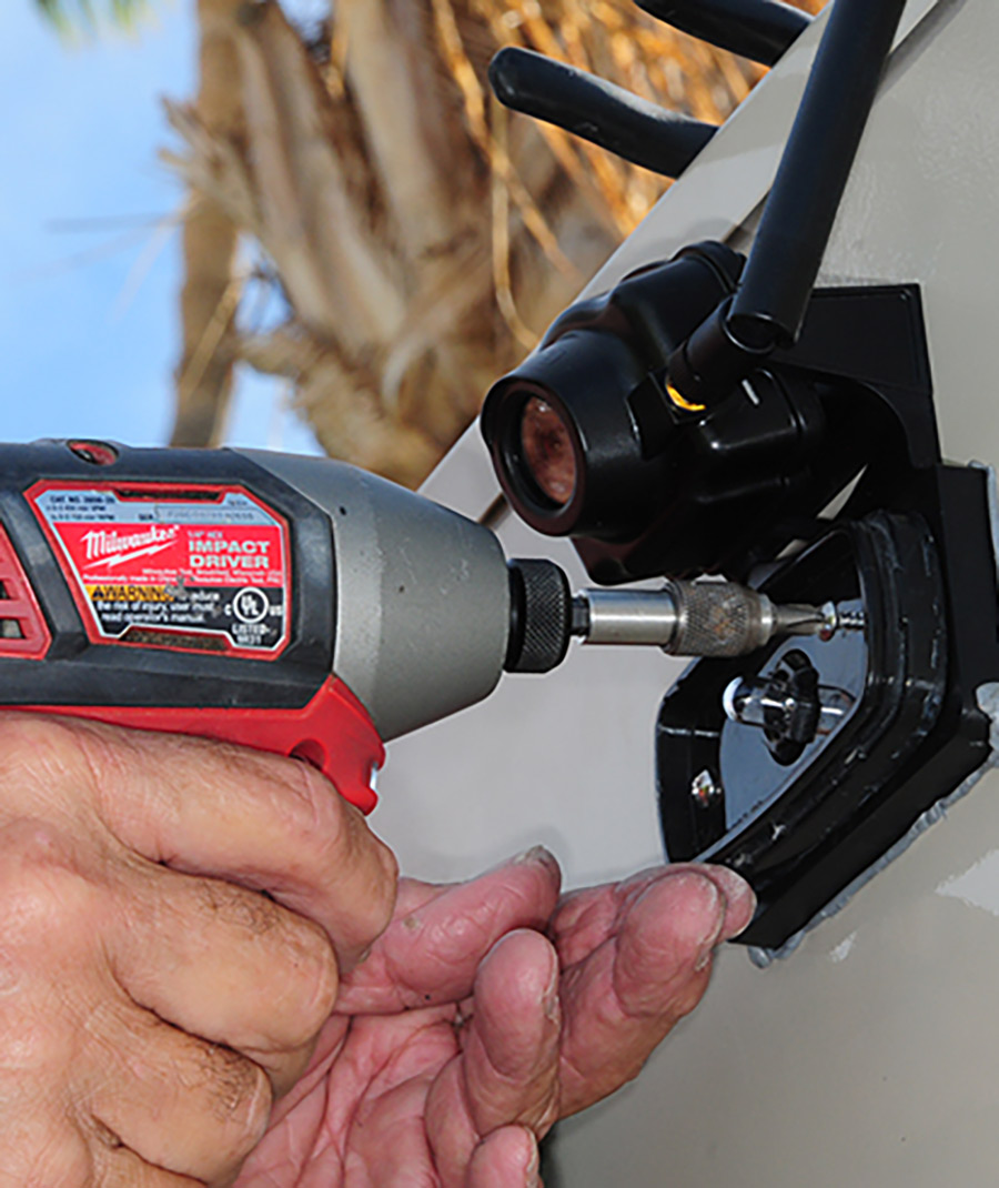 Portrait photograph close-up view of Bill Gehr's hands using a screw gun to firmly secure the bracket to the wall via provided stainless-steel screws on the back surface area of a RV trailer